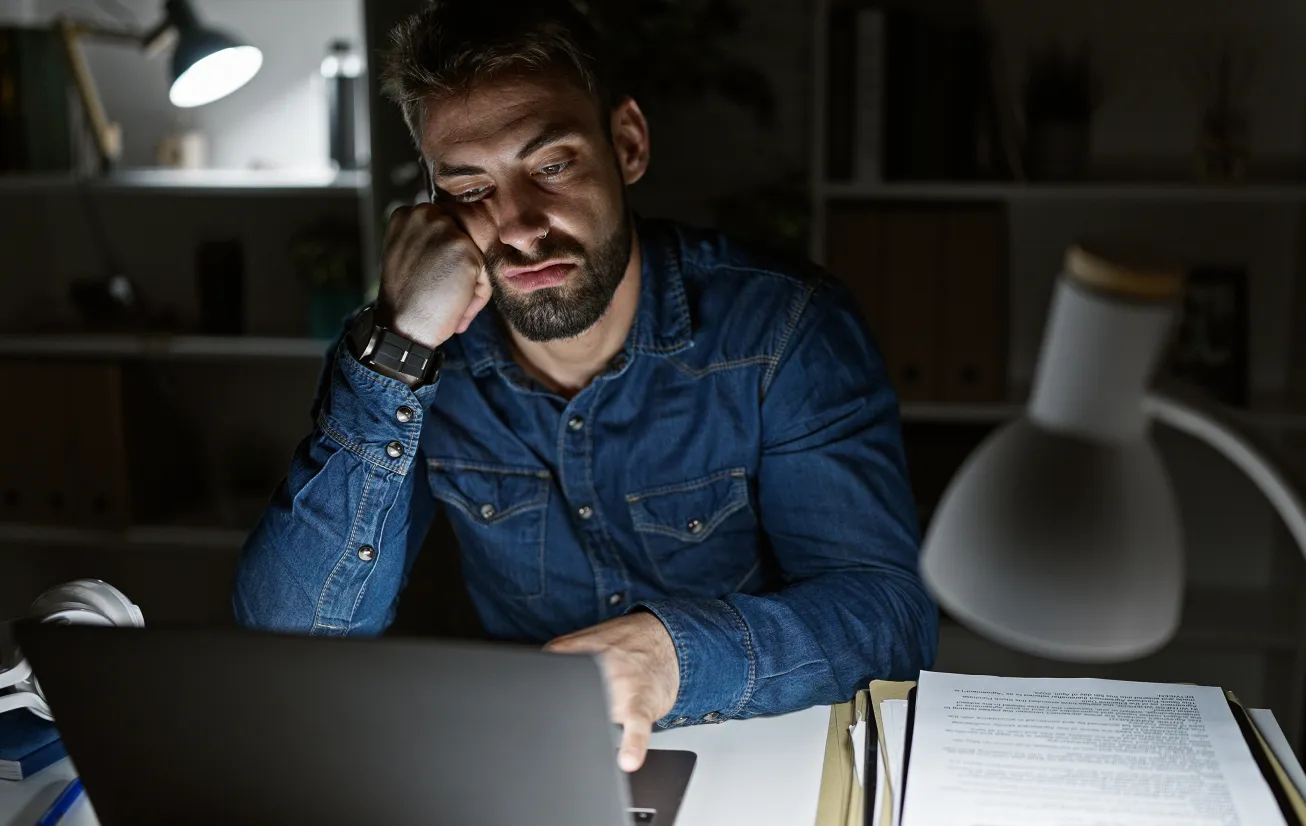 Man in denim shirt looking tired and resting his head on his hand while working late on a laptop at a desk with documents and a desk lamp.